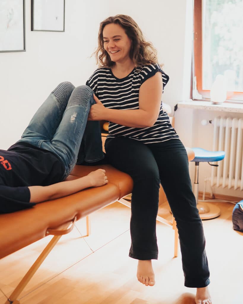 Claudia Hannemann, Mastertrainerin für Reflexintegration und Begründerin der AART Reflexintegration, sitzt barfuß auf einer Behandlungsliege und führt eine Reflexintegrationsübung mit einem Kind durch. Sie hält dessen Knie in einer ruhigen, achtsamen Position. Die Situation zeigt eine typische Sequenz aus dem Reflexintegrationstraining.