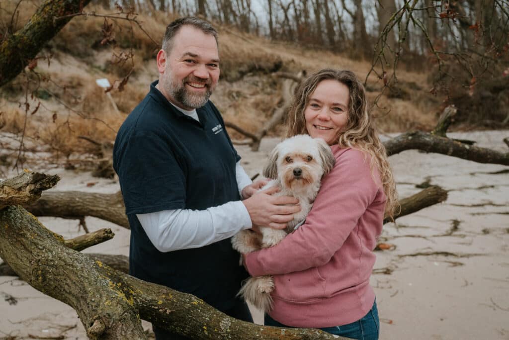 Claudia Hannemann und Sören Doll, Gründer der Awesome Academy stehen gemeinsam und schauen in die Kamera. Claudia Hannemann hält den Schul- und Therapiehund Cappuccino auf dem Arm. Sie stehen am Strand, im Hintergrund sind Dünen sichtbar und ein alter Baumstumpf.