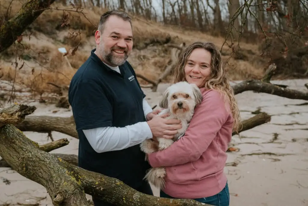 Claudia Hannemann und Sören Doll, Gründer der Awesome Academy stehen gemeinsam und schauen in die Kamera. Claudia Hannemann hält den Schul- und Therapiehund Cappuccino auf dem Arm. Sie stehen am Strand, im Hintergrund sind Dünen sichtbar und ein alter Baumstumpf.