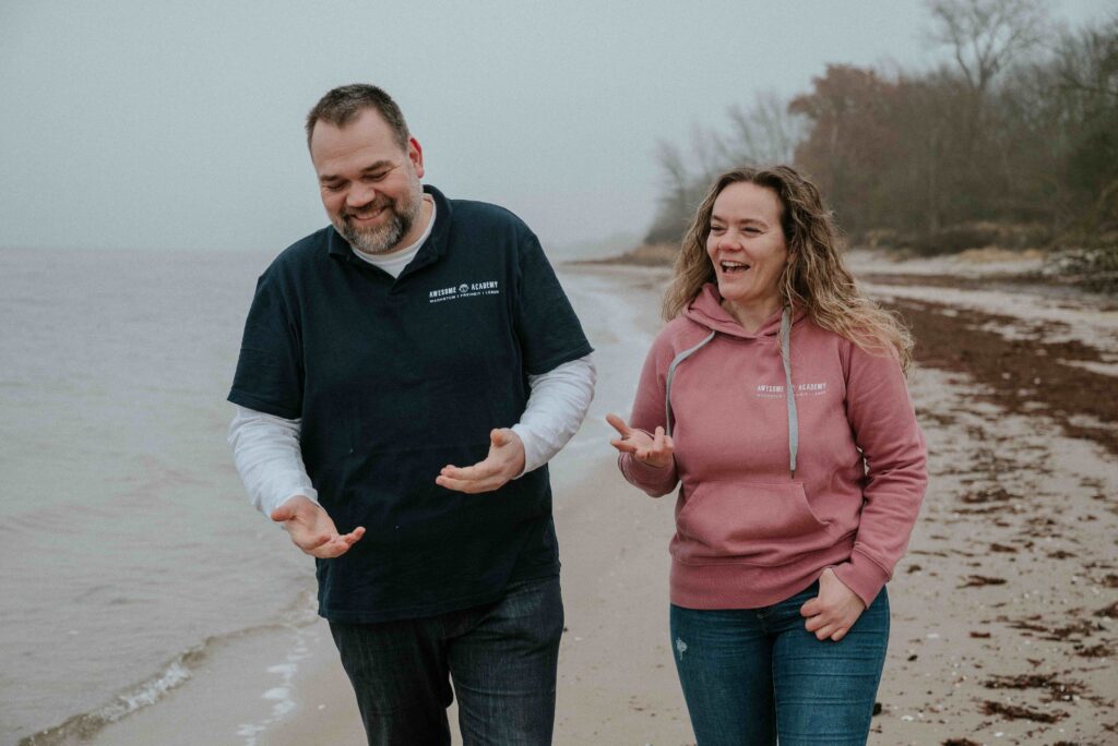 Zwei Menschen, lächelnd und im Gespräch, spazieren gemeinsam am Strand entlang. Beide tragen Kleidung mit dem Logo der Awesome Academy. Die Szene wirkt offen, wertschätzend und kollegial.