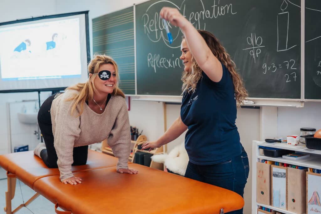 Zwei Frauen bei einer Reflexintegrationseinheit: Eine befindet sich im Vierfüßlerstand mit Augenklappe, die andere führt eine gezielte visuelle Reizung mit einem Objekt durch.
Claudia Hannemann, Gründerin der Awesome Academy, leitet das Reflexintegrationstraining an.