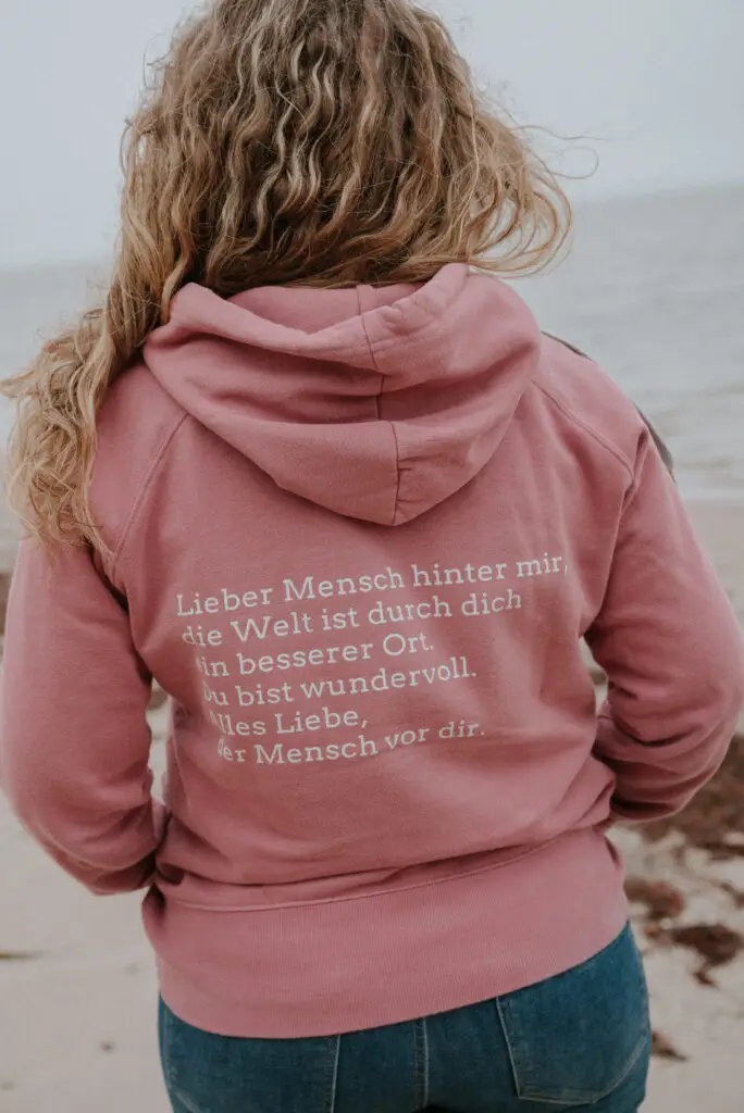 Claudia Hannemann am Strand mit Hoodie-Botschaft: Lieber Mensch hinter mir, die Welt ist durch dich ein besserer Ort – Wertschätzung für Fachkräfte in der Reflexintegration